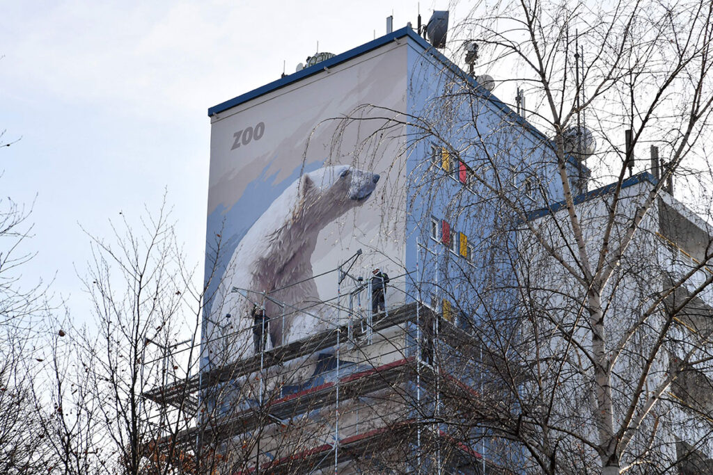 Wandmalerei und Fassadengestaltung bei diesem Wandgemälde eines Eisbären in der Ziolkowskistraße 9 in Rostock entstand nach eigener Idee. Ich konnte sowohl den ZOO als Begünstigten als auch die WIRO als Finanzierer dieser Idee gewinnen. Die Malerei ist ca. 20 m hoch und mehr als 8 m breit. Die Ausführungszeit lag von September bis Dezember 2016. Das Bärchen ist weithin zu sehen, schließlich befindet sie sich an einem Gebäude, das mehr als 40 m hoch ist.